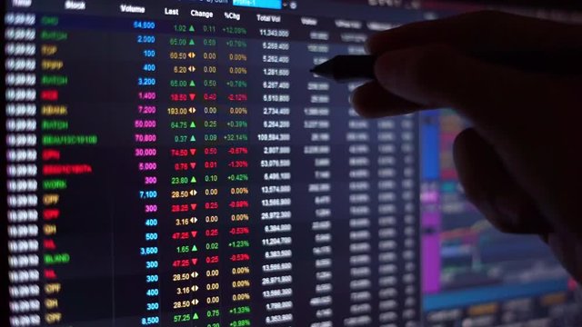 financial chart with moving hand holding pen finding number in stock market ticker board on black monitor background