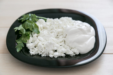 Black glass plate with cottage cheese and sour cream on a wooden background. Cottage cheese with sour cream on a black plate.