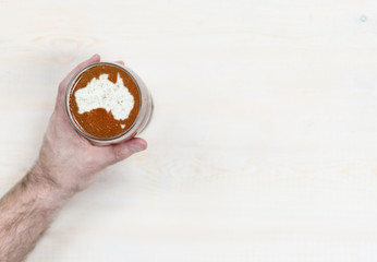 Fototapeta na wymiar Male hand holding mug of beer with silhouettes of Australia on foam. Top view. Space for text