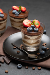 Classic tiramisu dessert with blueberries and strawberries in a glass on stone serving board on dark concrete background
