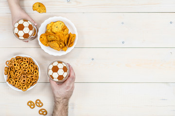 Friends holds glass mug of beer with a soccer ball on a beer foam and pretzels with chips on light wooden background. Empty space for text. Top view