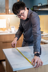 Asian man designer using tape measure for measuring white granite countertops on modern kitchen counter in showroom. Shopping furniture material for home improvement. Interior design concept