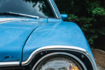 Close up of vintage blue car's old headlamp. Parked and surrounded by trees.