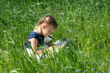 Little girl with a book in the garden. Kid is readding a book. A little girl 4-5 years old sits on the grass and reads a book.