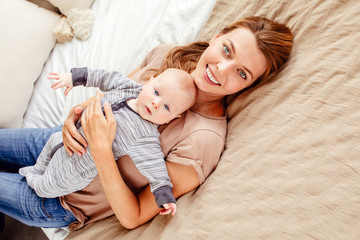 Beautiful young woman with cute baby smiling and looking at camera while lying on comfortable bed at home