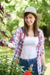 Beautiful smiling Caucasian brunette with hat, gardening gloves and in working clothes taking care of her flowers in backyard.