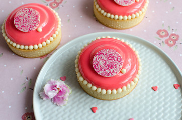Mousse mini tarts covered with pink glaze and decorated with chocolate elements on white plate on pink background