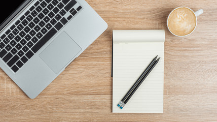 On the desktop, a laptop, an open notebook with pencils and a cup of coffee. View from above.