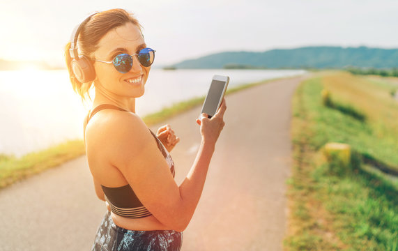 Young beautiful female starting jogging and listening to music using smartphone and  wireless headphones cheerfully smiling in camera