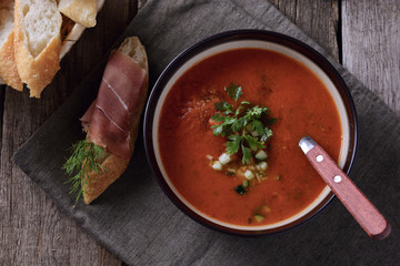 Cold tomato soup gazpacho in a bowl next to sandwich with ​​jamon on top are on a napkin on old wooden table, top view