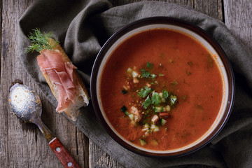 Cold tomato soup gazpacho in a bowl next to sandwich with ​​jamon on top are on a napkin and spoon on old wooden table, top view