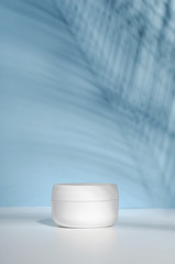 A white jar of cream on a stand on a blue background, on a white table with tropical palm leaves and their shadow. Stylish look of the product, mock up, identity.