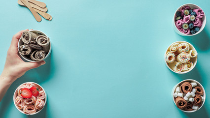 Hand with rolled ice cream in cone cup on blue background. Different iced rolls top view or flat...