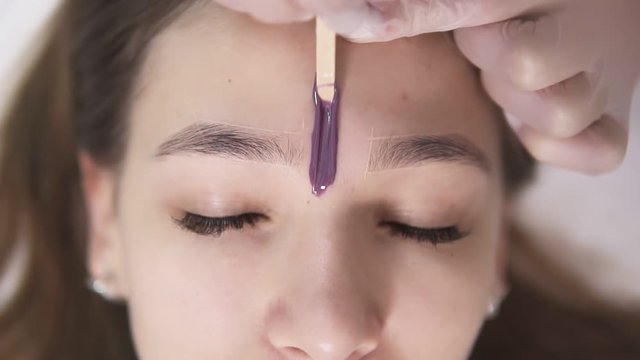 The client is lying on couch during eyebrowes treatment at studio beauty, beautician depilating and shaping brows at beauty salon, applying a bit of wax between the brows. Above head footage. Slow