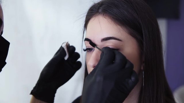 A beautician in black gloves using a white clean cotton disk ank little brows brush to remove the paint from the brows A woman sits in the chair during a visit at the beauty salon. Slow motion