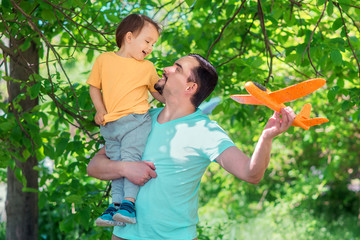 Father and toddler son playing together with orange toy plane outdoors: boy is sitting on shoulder of man, both dad and kid are looking at each other and laughing. Happy family relationships concept