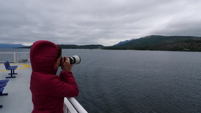 Tourist woman on windy cruise ship deck enjoying fjord view, taking photo with camera. Ferry ride route Bognes - Lodingen to Lofoten islands Norway. Tourism vacation and travel