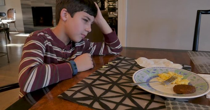 Unhappy young 8 - 10 year old boy sitting at a table not eating his breakfast