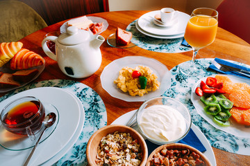 Breakfast table with variety of foods including cereals, yoghurt, scrambled eggs, fruit, croissant and drinks such as tea, coffee and orange juice