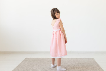 The little charming girl in a pink dress smiles and looks into the camera against the background of a white wall. Concept of the author's children's clothing. Copy space