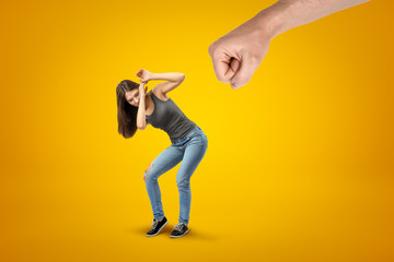 Fototapeta na wymiar Young brunette girl wearing casual jeans and t-shirt protecting herself from big male stretched fist on yellow background