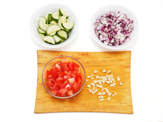 Sliced tomato in a transparent bowl and chopped garlic on a cutting board. White background. Sliced red onions and cucumbers in a white bowls