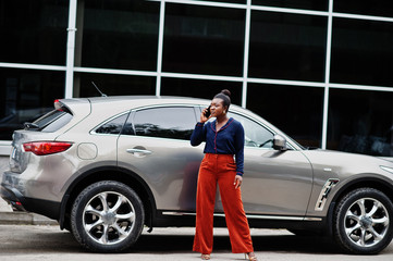Rich business african woman in orange pants and blue shirt posed against silver suv car and speak...