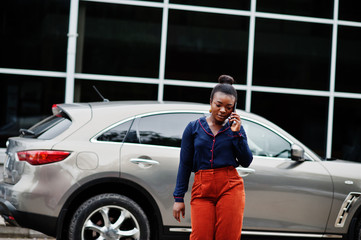 Fototapeta na wymiar Rich business african woman in orange pants and blue shirt posed against silver suv car and speak on mobile phone.
