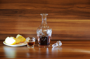 An open carafe of whiskey, a stack of whiskey and pieces of sliced apple on a saucer on a wooden...