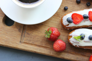 A Cup of hot black coffee and delicious eclairs with strawberries and blueberries on a wooden cutting Board. Copy space. Beautiful and delicious dessert.