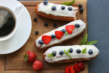 A Cup of hot black coffee and delicious eclairs with strawberries and blueberries on a wooden cutting Board. Copy space. Beautiful and delicious dessert.