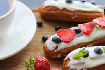 A Cup of hot black coffee and delicious eclairs with strawberries and blueberries on a wooden cutting Board. Copy space. Beautiful and delicious dessert.