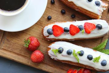 A Cup of hot black coffee and delicious eclairs with strawberries and blueberries on a wooden cutting Board. Copy space. Beautiful and delicious dessert.