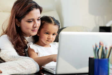 Young mother sitting at the table together with her little daughter and they typing on laptop computer at home