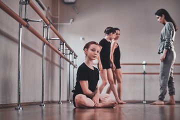 Obraz premium Little pretty ballerina is sitting on the floor while her other classmates doing exercises with trainer.