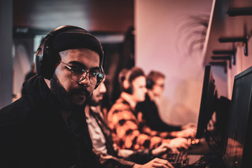 Portrait of a gamer with hat, glasses and headphones with other teamplayers at background.