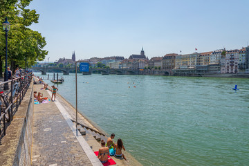 Fototapeta na wymiar Outdoor scenery of people enjoy, relax and chill out on riverside and also swim and float on Rhine river during hot summer season with background of cityscape in Basel, Switzerland. 