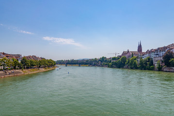 Obraz premium Outdoor sunny scenery of promenade and bridge along riverside, Basel's cityscape, Münster Cathedral and people float and swim, from the bridge cross Rhine river in Basel, Switzerland in summer season.