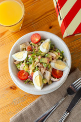 Top view of traditional american cobb salad with chopped avocado, tomatoes, hard boiled egg, roasted chicken and greens for a healthy lunch, served on wooden table with soft light