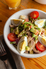 Traditional american cobb salad with chopped avocado, tomatoes, hard boiled egg, roasted chicken and greens for a healthy lunch, served on wooden table with soft light
