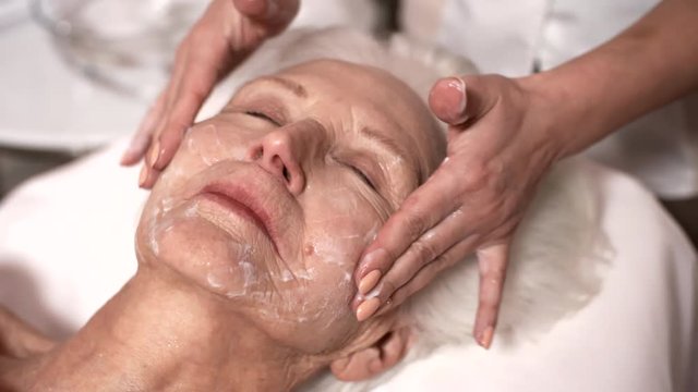 Close-up shot of beautiful senior Caucasian lady lying on couch in beauty parlour, and female beautician gently patting cream over her face, then giving her face massage with delicate hand movements