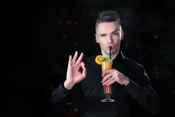 A handsome guy in a black shirt is holding a cocktail of orange juice in his hand and showing a hand gesture. Portrait of a guy of European appearance on a black background. Copy space.