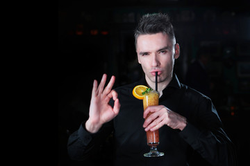 Fototapeta premium A handsome guy in a black shirt is holding a cocktail of orange juice in his hand and showing a hand gesture. Portrait of a guy of European appearance on a black background. Copy space.