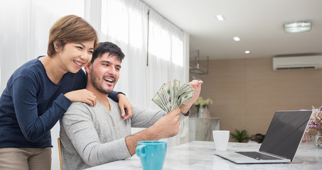 Portrait of young happy couple man woman at desk counting lot of money with computer, hands holding...