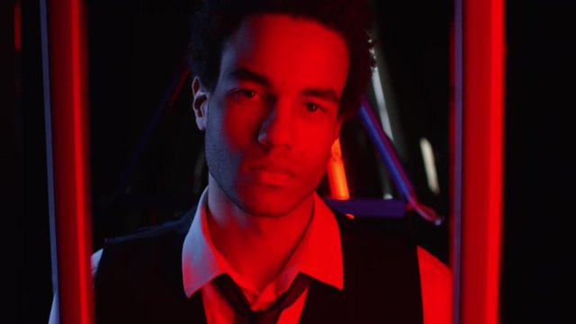 Tilt up of young mixed-race man illuminated in red light raising his head and looking at camera on black background with colorful neon lamps