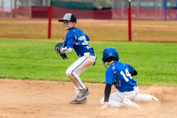 Youth baseball player in blue uniform playing short stop avoiding the sliding base runner and preparing to throw the ball.