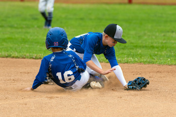 Youth baseball player in blue uniform playing short stop withstanding base runner sliding into the...