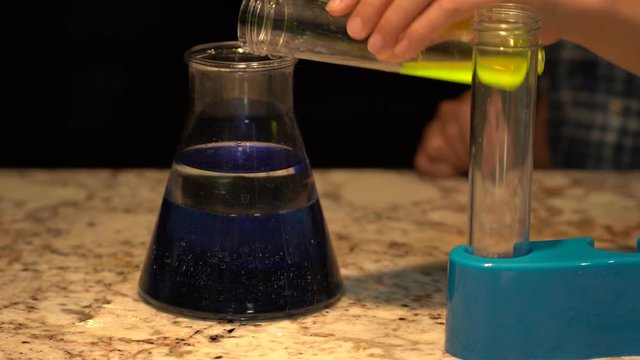 Small hand pouring a fluorescent green liquid substance out of a plastic test tube into a beaker cup containing blue water and baby oil. He then turns off the light and you can see the beaker glow.