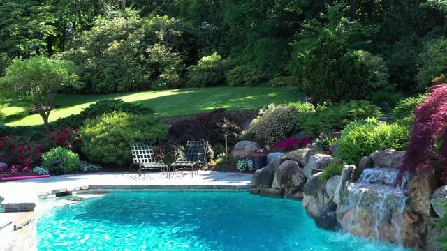 a slow dolly shot over a beautiful summer pool setting. A steady drone flight towards pool's waterfall on the right and background green and luscious lawn in the back