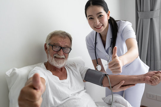 senior man with smiling nurse, takes care and discussion after health check by thumb up cheer on bed at nursing home